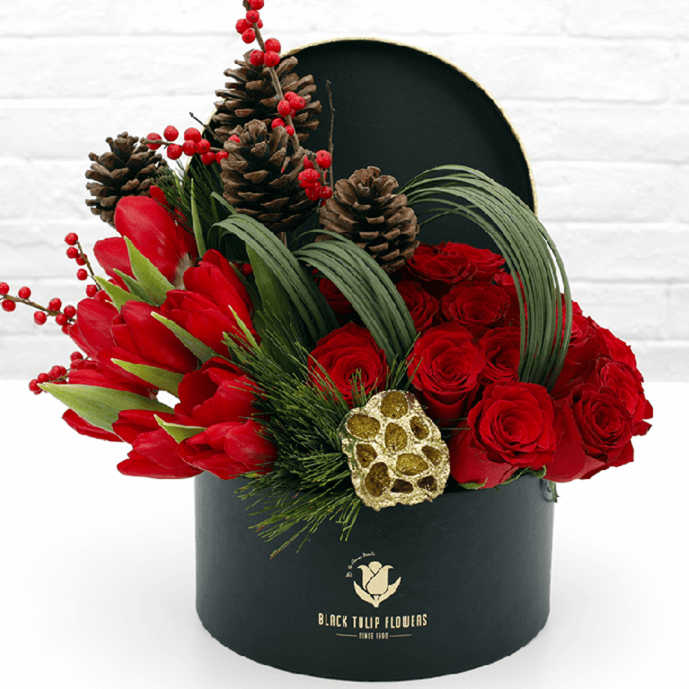 Red exotic flowers and roses with pinecones, berries and greenery arranged in a black round box