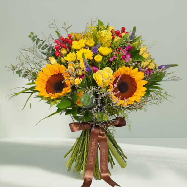 Hand-tied bouquet with fresh sunflowers, roses, spray roses, chrysanthemums, veronica, berries, rescues, and eucalyptus finished with a brown ribbon.