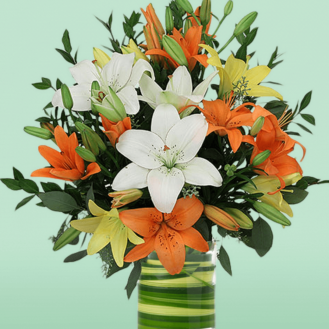 A transparent glass cylinder vase filled with orange, white and yellow lilies with fresh greenery.