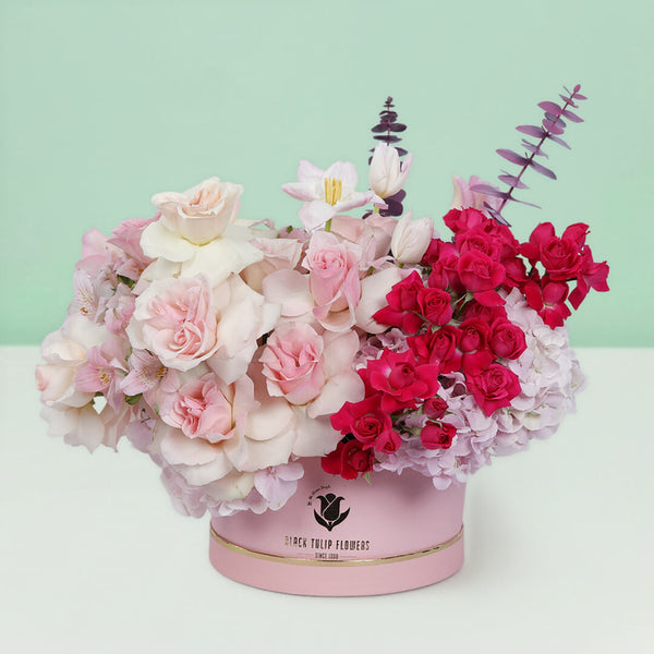 Roses, Spray Roses, Tulips and Hydrangeas with greenery arranged in a pink round flower box.