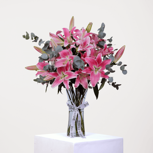 Vase arrangement featuring pink lilies and eucalyptus greenery in a transparent cylinder glass vase, finished with a branded ribbon.