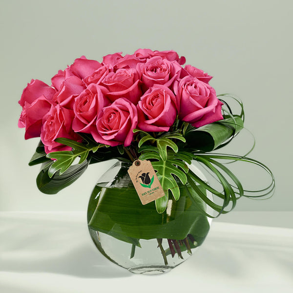 Pink roses and decorative tropical greenery in a transparent fish bowl glass vase.