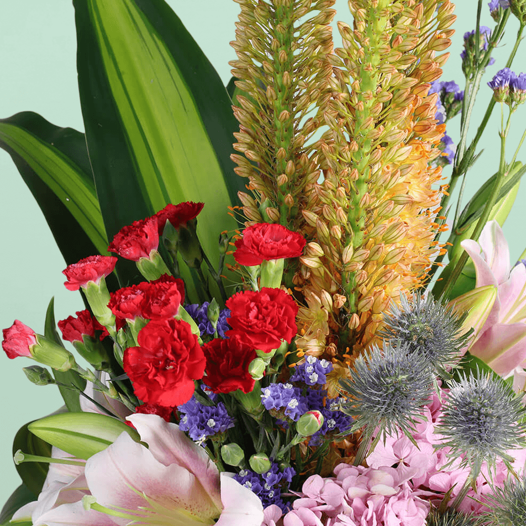 A square glass vase filled with eremurus, hydrangea, lilies, spray carnations, eryngium and statice.