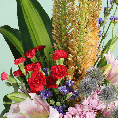 A square glass vase filled with eremurus, hydrangea, lilies, spray carnations, eryngium and statice.