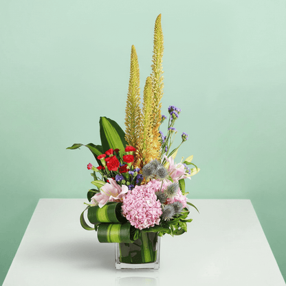 A square glass vase filled with eremurus, hydrangea, lilies, spray carnations, eryngium and statice.
