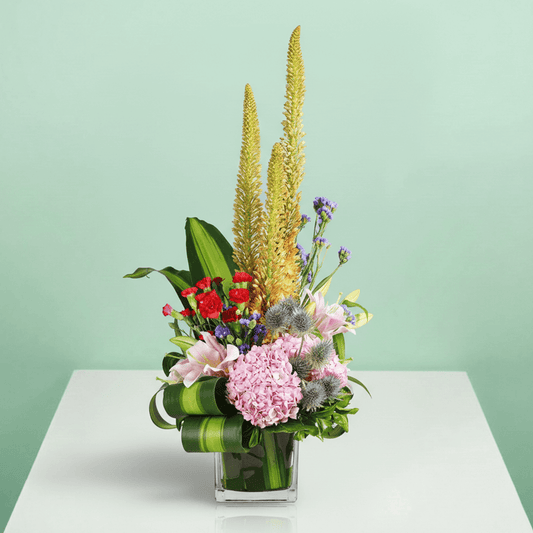 A square glass vase filled with eremurus, hydrangea, lilies, spray carnations, eryngium and statice.