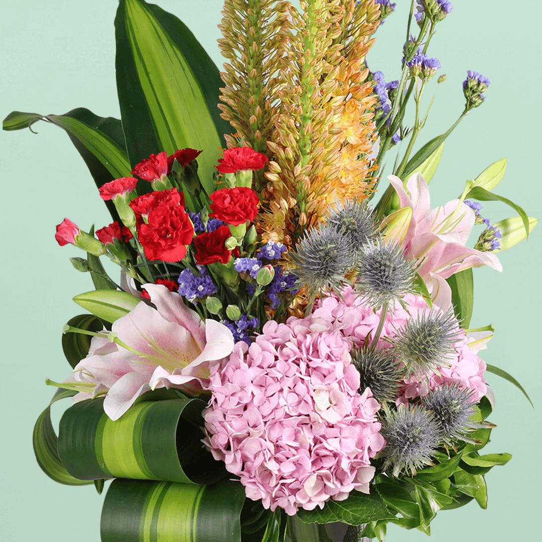 A square glass vase filled with eremurus, hydrangea, lilies, spray carnations, eryngium and statice.