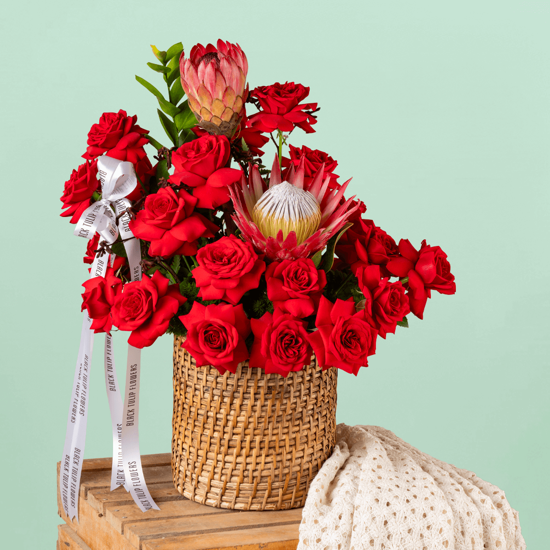 Red roses and protea arranged with greenery in a woven round basket