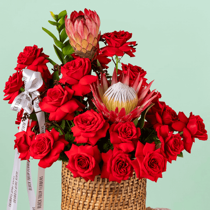 Red roses and protea arranged with greenery in a woven round basket