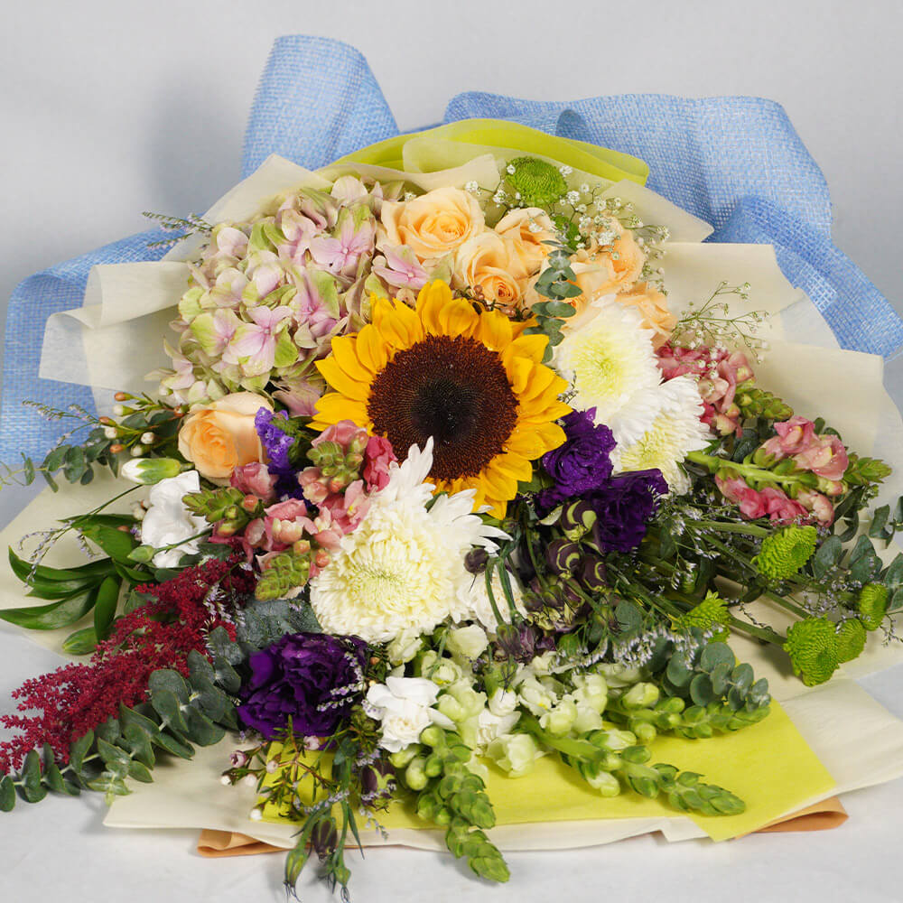 A cheerful bouquet of assorted blooms featuring Sunflower, Hydrangea, Roses, Snapdragon, Eustoma, Astilbe, Chrysanthemum and Wax Flower. Fresh greenery and soft blue non-woven wrapping create a bright, garden-inspired presentation. A thoughtful choice for birthdays, congratulations, thank-you gifts and joyful celebrations.