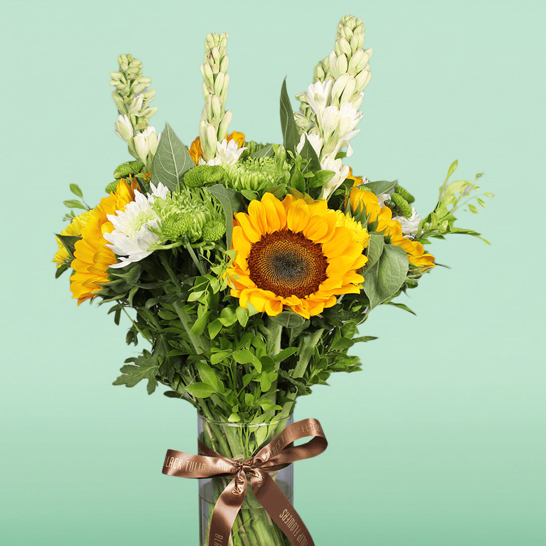 Yellow sunflowers, white tuberose, green disbuds and chrysanthemums arranged with fresh greenery in a glass vase