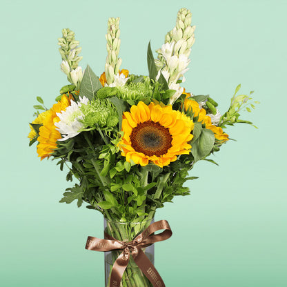 Yellow sunflowers, white tuberose, green disbuds and chrysanthemums arranged with fresh greenery in a glass vase