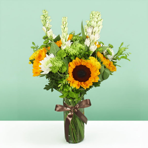 Yellow sunflowers, white tuberose, green disbuds and chrysanthemums arranged with fresh greenery in a glass vase