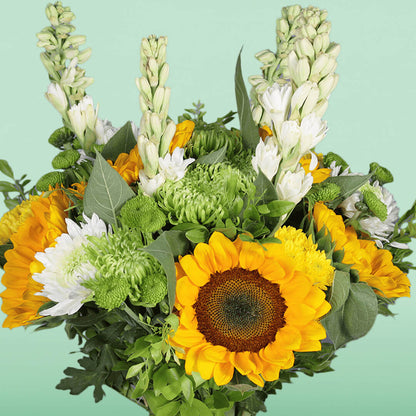 Yellow sunflowers, white tuberose, green disbuds and chrysanthemums arranged with fresh greenery in a glass vase