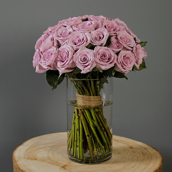 Fresh purple roses and vibrant foliage are beautifully styled in a transparent cylinder glass vase