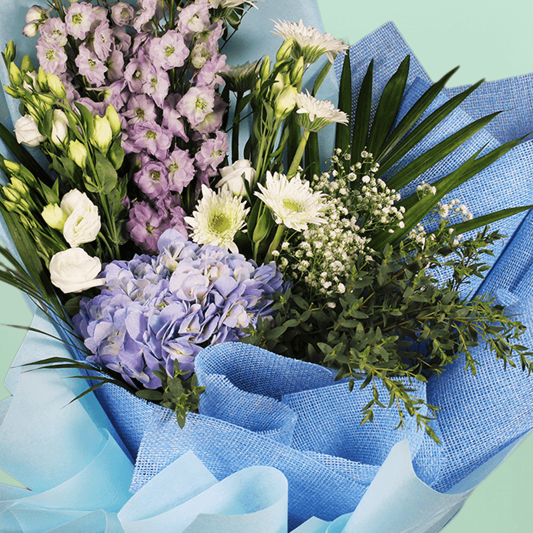 A bouquet of blue hydrangea, white chrysanthemums, Eustoma and delphinium with fresh greenery wrapped in blue non-woven paper.
