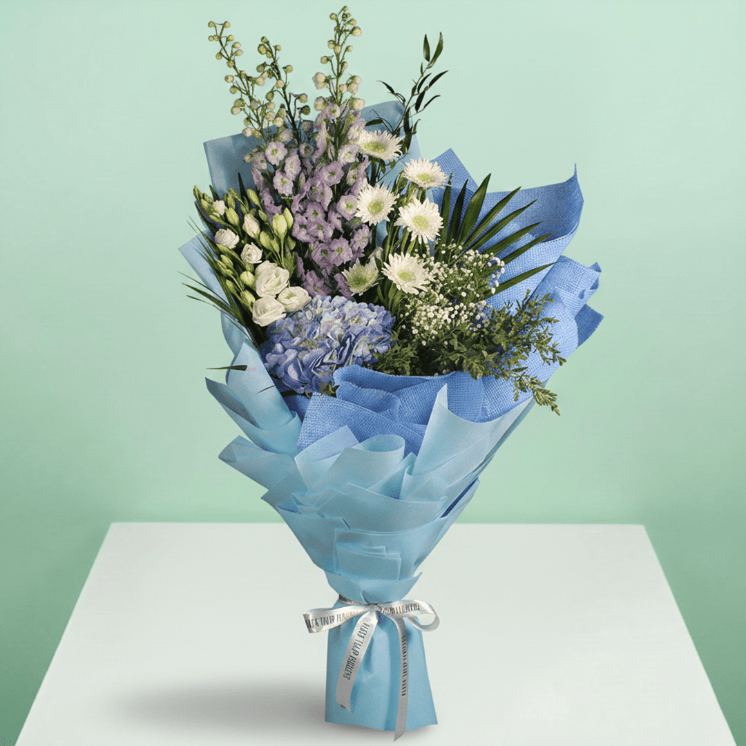 A bouquet of blue hydrangea, white chrysanthemums, Eustoma and delphinium with fresh greenery wrapped in blue non-woven paper.
