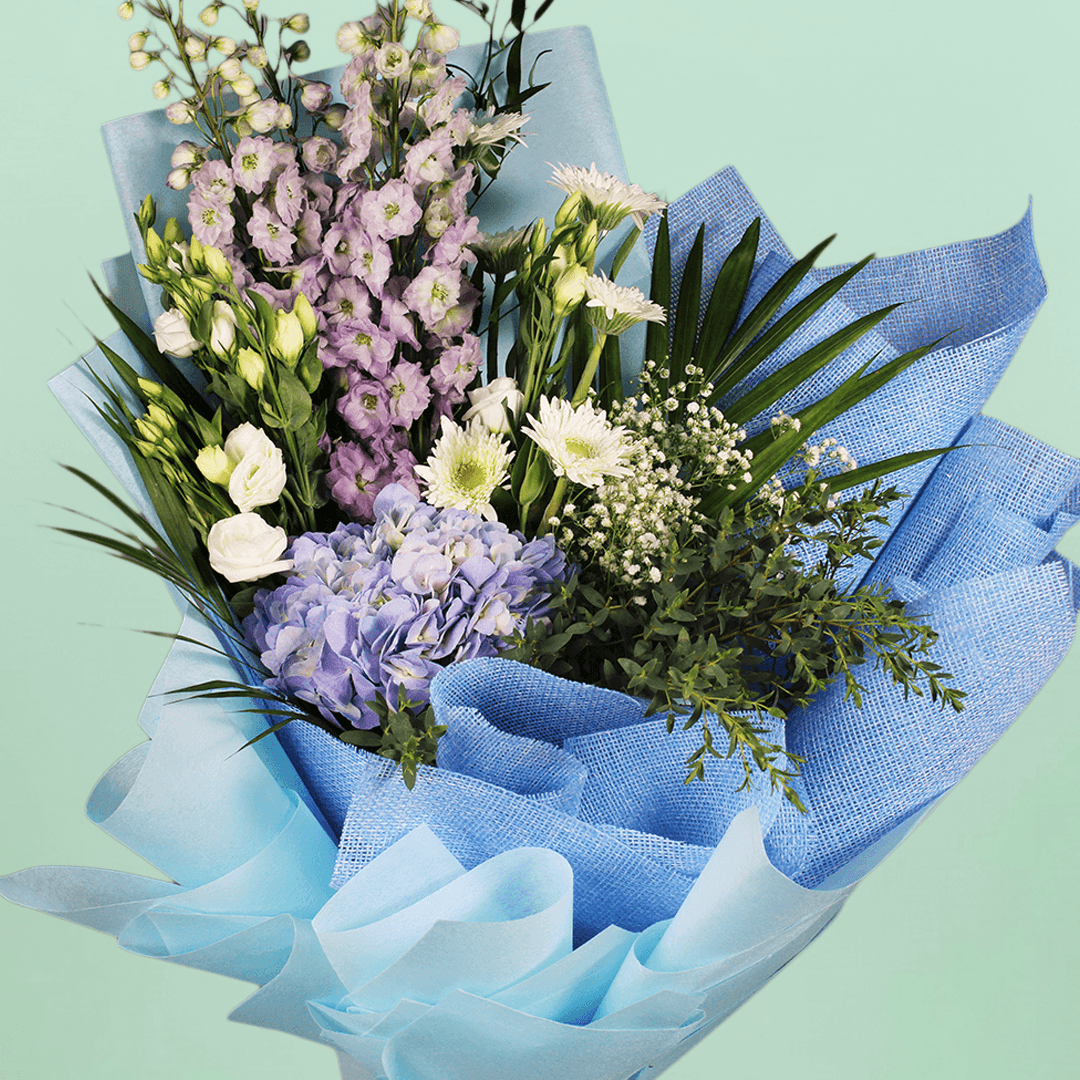 A bouquet of blue hydrangea, white chrysanthemums, Eustoma and delphinium with fresh greenery wrapped in blue non-woven paper.
