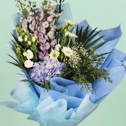 A bouquet of blue hydrangea, white chrysanthemums, Eustoma and delphinium with fresh greenery wrapped in blue non-woven paper.
