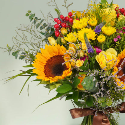 Hand-tied bouquet with fresh sunflowers, roses, spray roses, chrysanthemums, veronica, berries, rescues, and eucalyptus finished with a brown ribbon.
