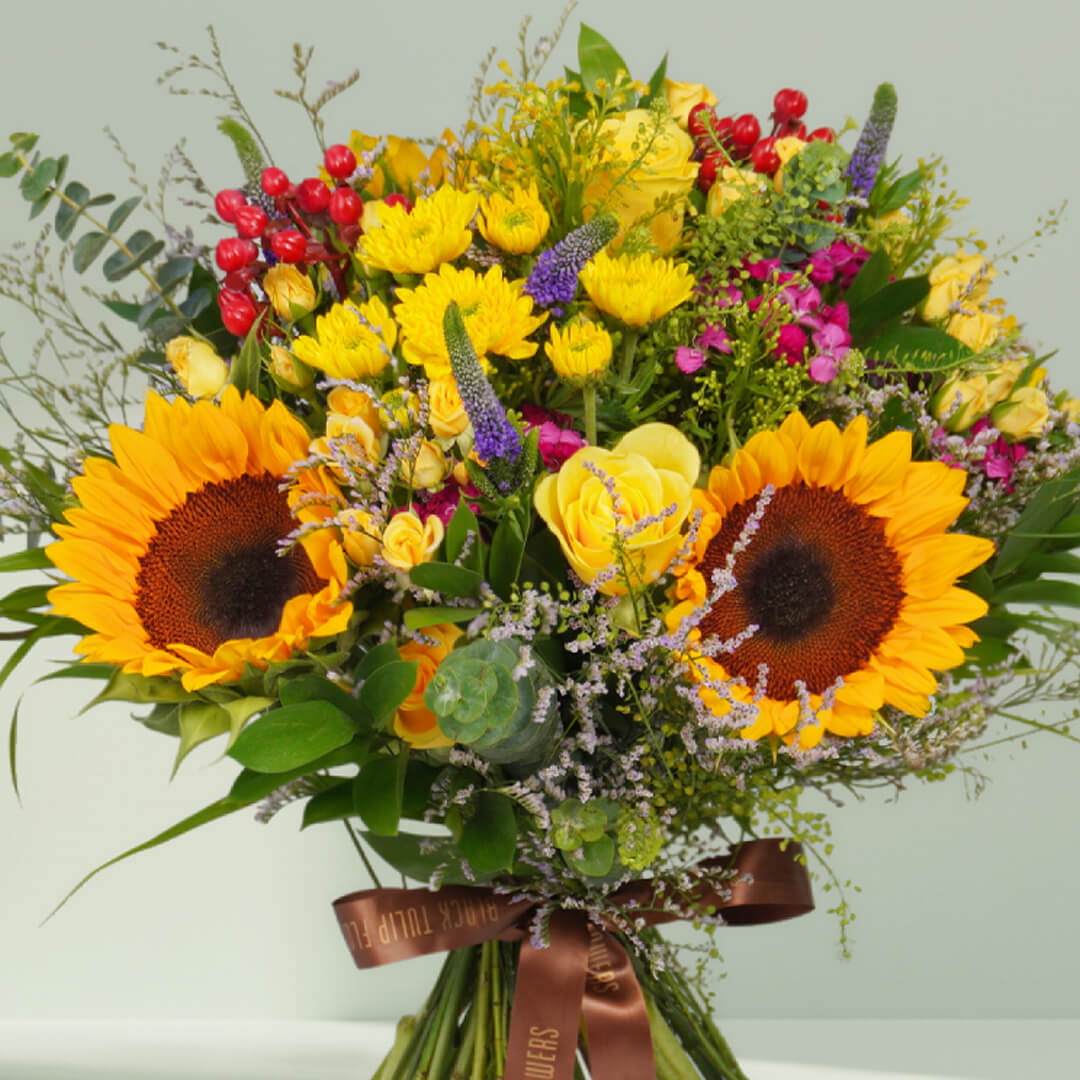 Hand-tied bouquet with fresh sunflowers, roses, spray roses, chrysanthemums, veronica, berries, rescues, and eucalyptus finished with a brown ribbon.