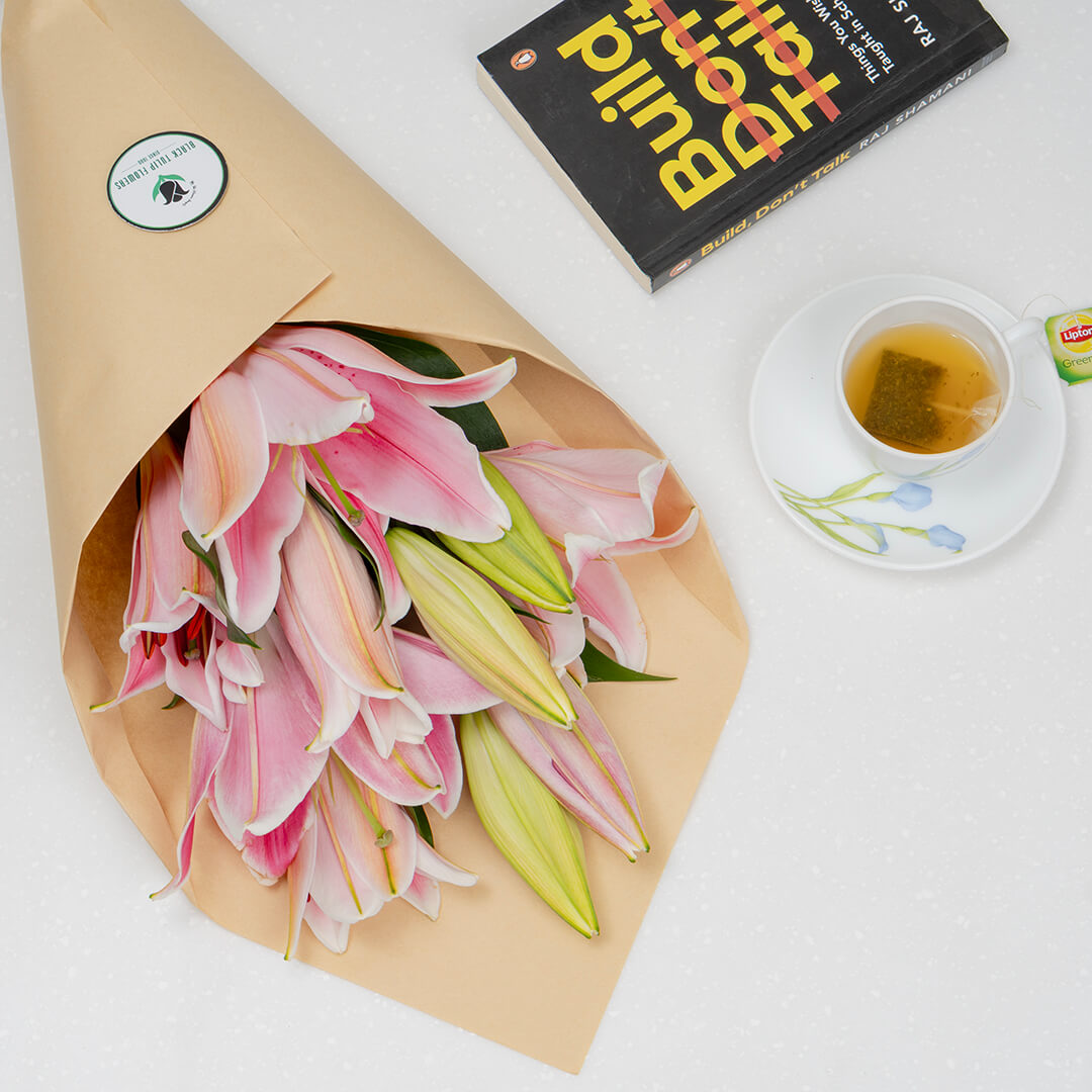 A bouquet of pink and green lilies wrapped in brown kraft paper, placed on a white surface next to a black book and a cup of tea on a saucer.