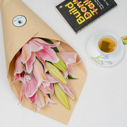 A bouquet of pink and green lilies wrapped in brown kraft paper, placed on a white surface next to a black book and a cup of tea on a saucer.