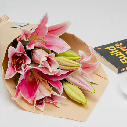 A bouquet of pink and white lilies with green buds, wrapped in brown paper, arranged on a white surface next to a book.