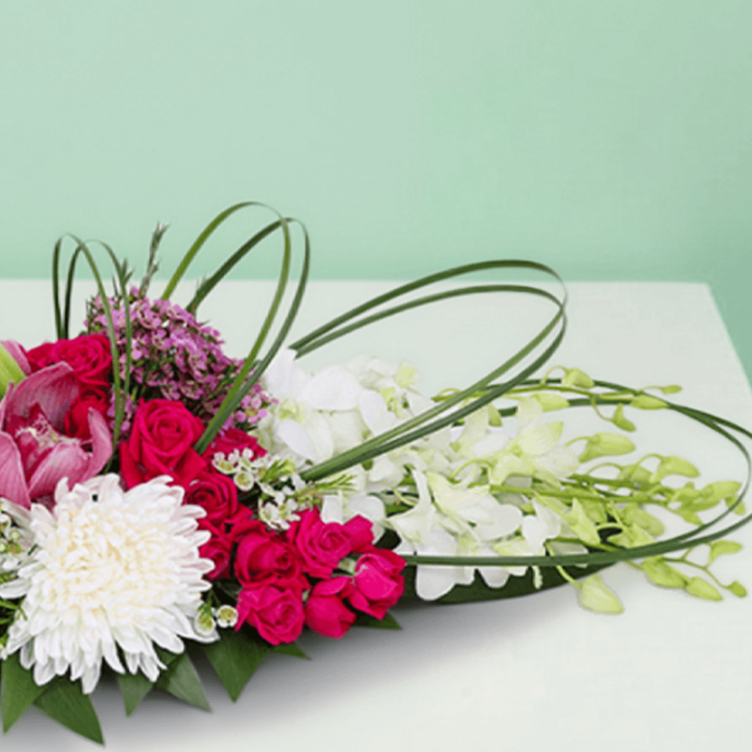 Long and low arrangement of orchids, lilies, roses, disbuds and chrysanthemums with curved greenery on a green tray