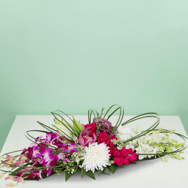 Long and low arrangement of orchids, lilies, roses, disbuds and chrysanthemums with curved greenery on a green tray