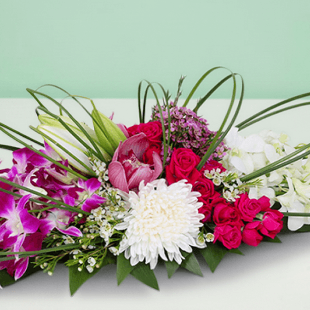 Long and low arrangement of orchids, lilies, roses, disbuds and chrysanthemums with curved greenery on a green tray