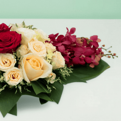 Long and low arrangement of yellow and burgundy orchids, cream and red roses, disbuds and greenery on a green tray