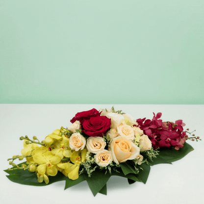 Long and low arrangement of yellow and burgundy orchids, cream and red roses, disbuds and greenery on a green tray