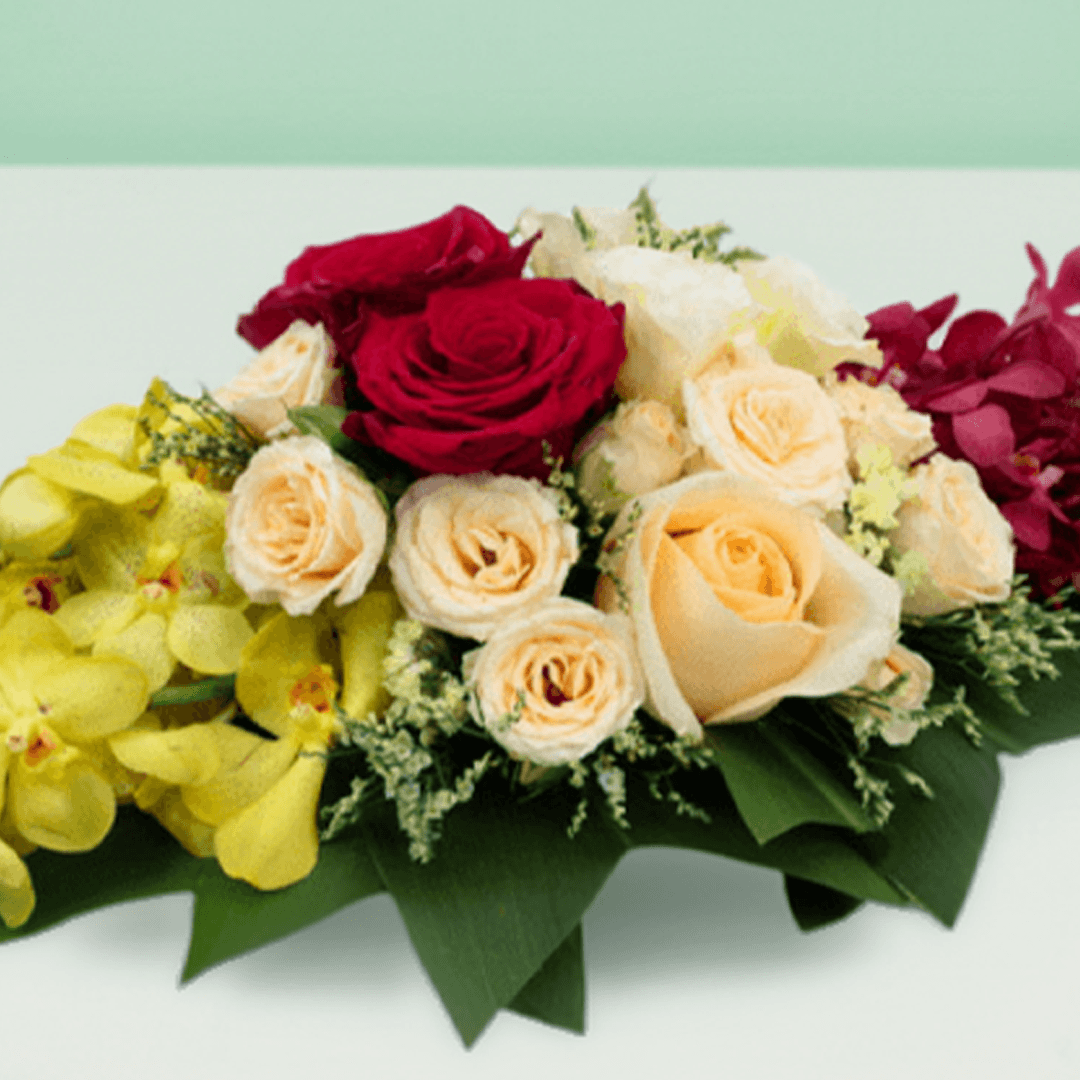 Long and low arrangement of yellow and burgundy orchids, cream and red roses, disbuds and greenery on a green tray