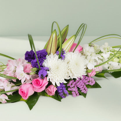Purple orchids, pink roses and white chrysanthemums arranged with curved greenery on a long green tray
