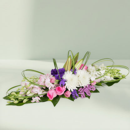 Purple orchids, pink roses and white chrysanthemums arranged with curved greenery on a long green tray

