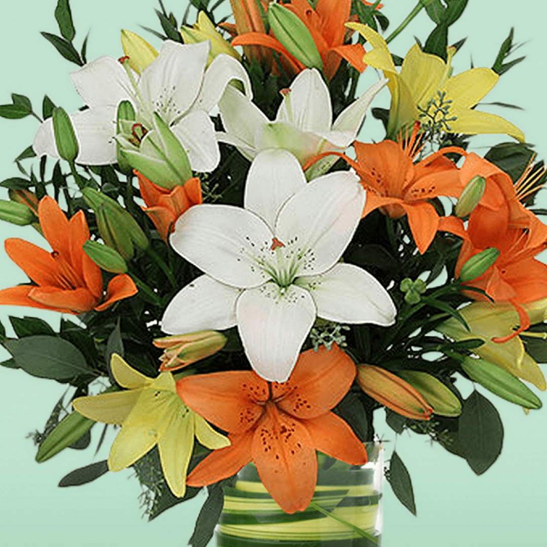 A transparent glass cylinder vase filled with orange, white and yellow lilies with fresh greenery.