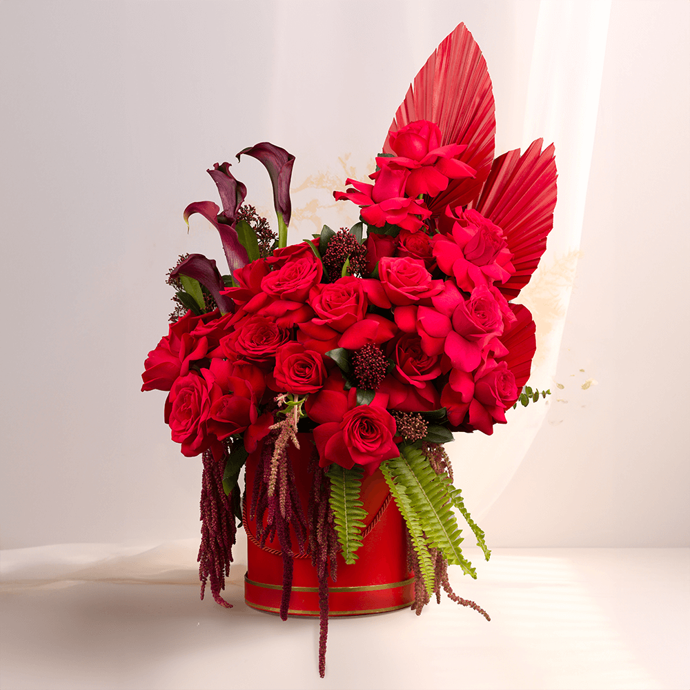 Red roses with Skimmia, cascading Amaranthus and dried palm leaves arranged in a red round flower box.