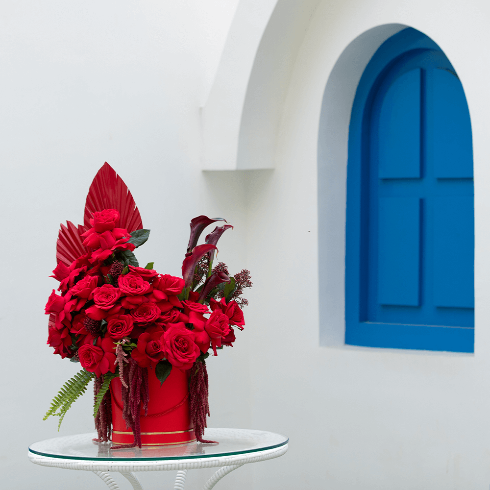Red roses with Skimmia, cascading Amaranthus and dried palm leaves arranged in a red round flower box.