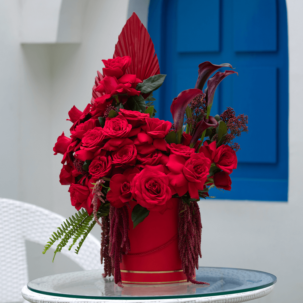 Red roses with Skimmia, cascading Amaranthus and dried palm leaves arranged in a red round flower box.