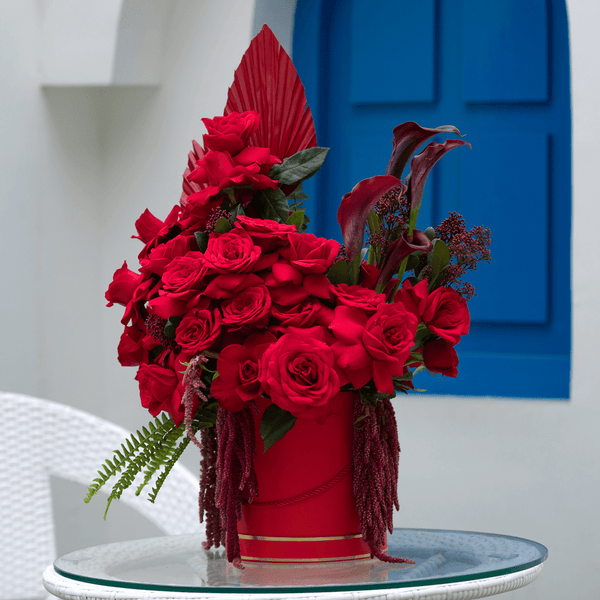 Red roses with Skimmia, cascading Amaranthus and dried palm leaves arranged in a red round flower box.
