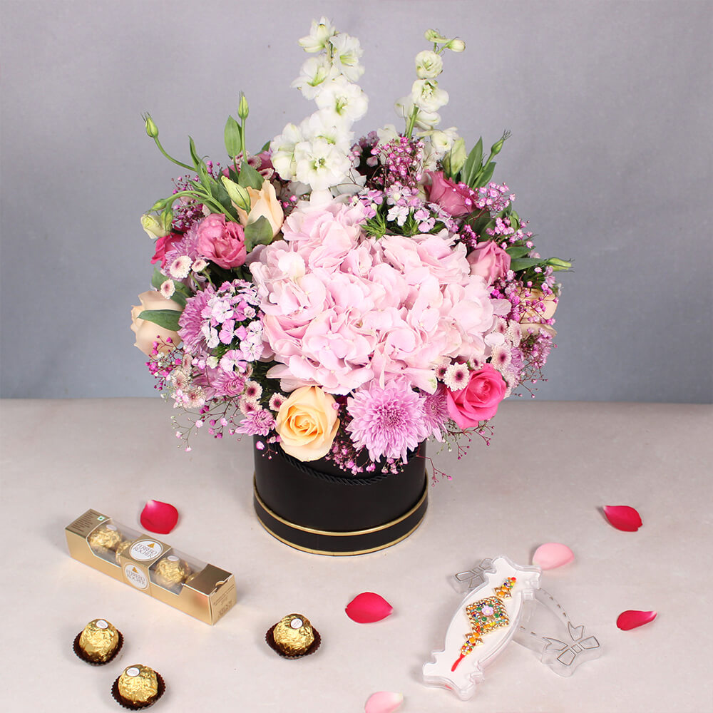 Pastel Hydrangeas, Roses, Delphiniums, Chrysanthemums and Eustoma in a black round box with Rakhi and Ferrero Rocher chocolates.
