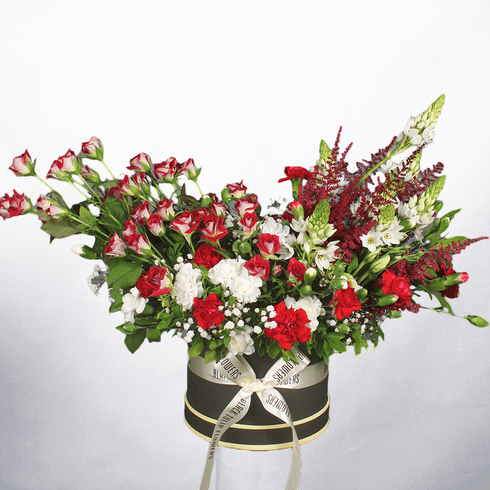Red spray roses, white carnations, astilbe and fresh greenery arranged in a black round flower box.
