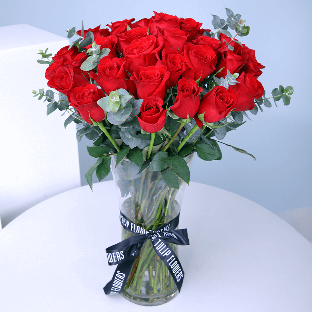 25 red roses with greenery arranged in a clear cylinder glass vase.
