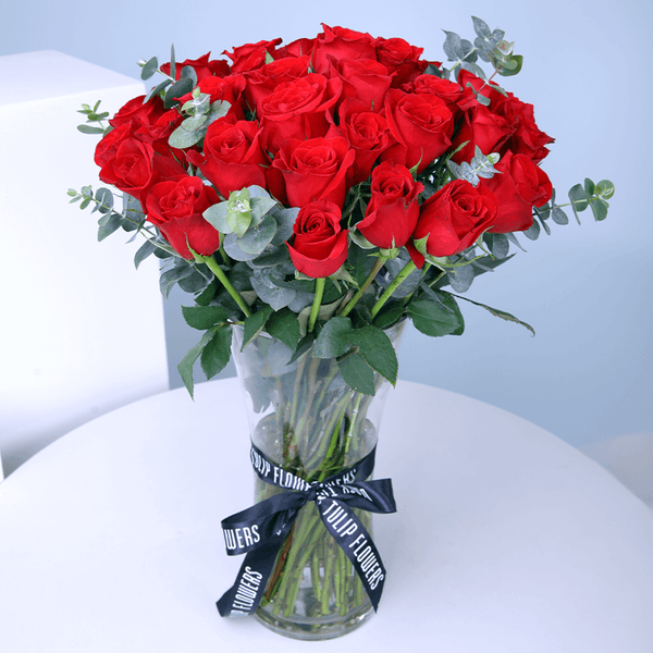 25 red roses with greenery arranged in a clear cylinder glass vase.
