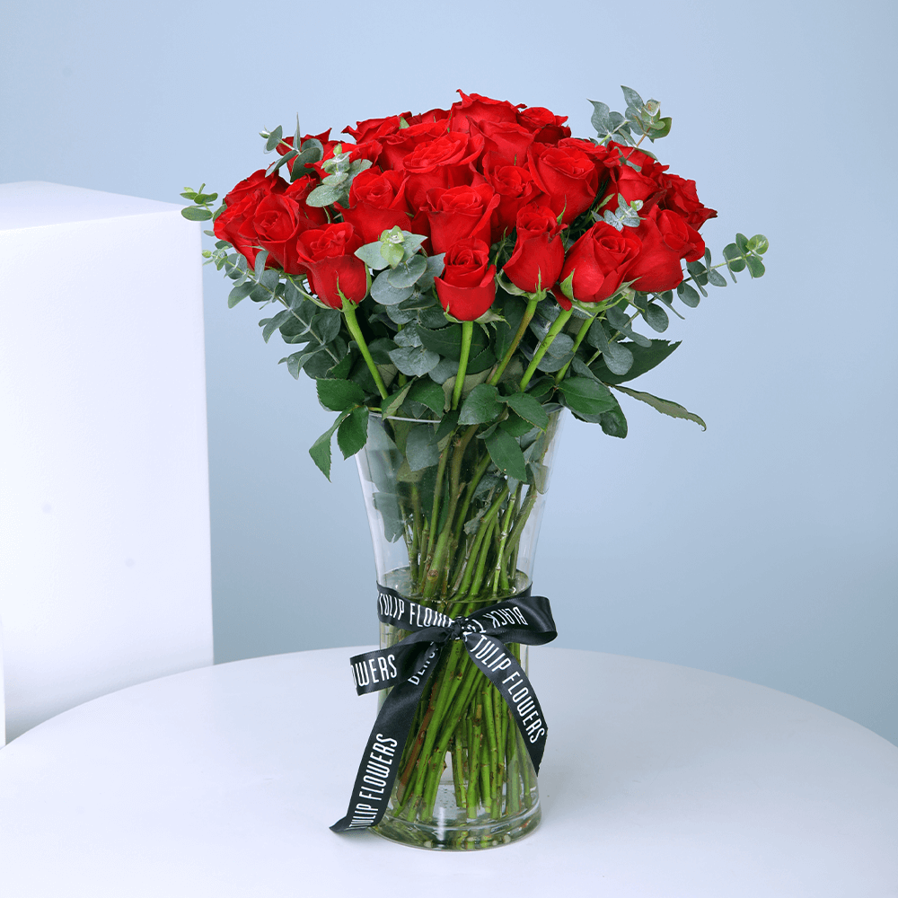 25 red roses with greenery arranged in a clear cylinder glass vase.
