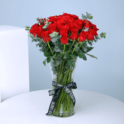 25 red roses with greenery arranged in a clear cylinder glass vase.