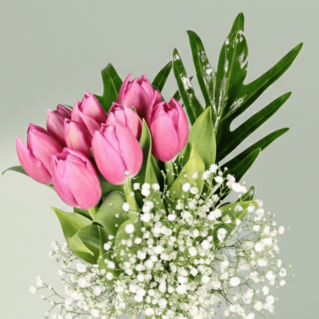 Pink tulip bouquet with baby’s breath and green foliage arranged in a clear glass vase.
