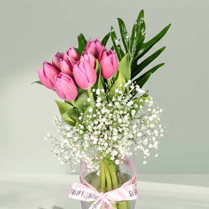 Pink tulip bouquet with baby’s breath and green foliage arranged in a clear glass vase.
