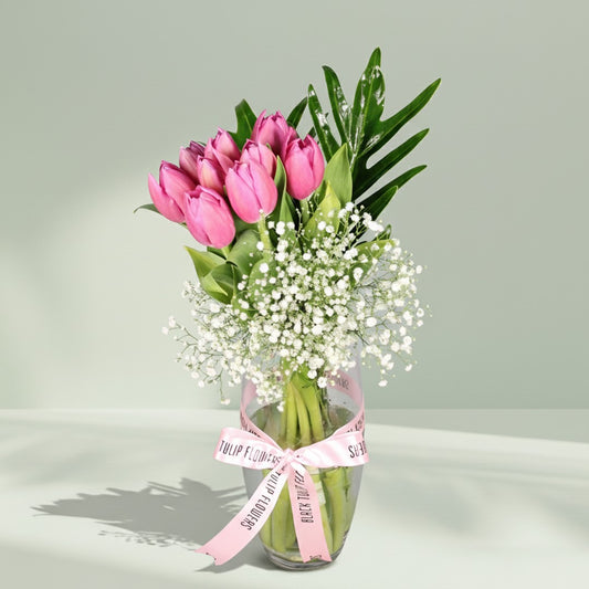 Pink tulip bouquet with baby’s breath and green foliage arranged in a clear glass vase.
